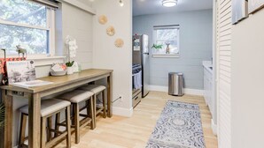A compact kitchen with a wooden table and bar stools, a refrigerator, a trash bin, and a patterned rug.