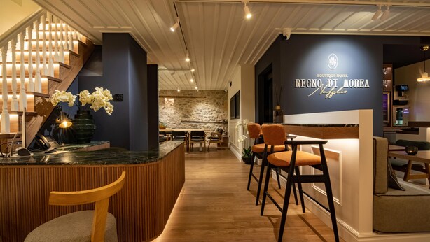 Un moderno vestíbulo de hotel con un mostrador de recepción de madera, taburetes de bar, una escalera y una pared de piedra al fondo.