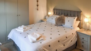 Un letto ben rifatto con una coperta a pois, due cuscini e asciugamani piegati. È presente un comodino con una lampada e una sveglia.