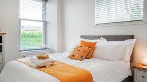 A neatly made bed with white bedding and an orange throw blanket, a bedside table with a lamp, and a window with blinds. - 2 bedroom accommodation in Falkland (Falkland)