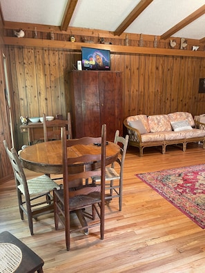 Um quarto com painéis de madeira, uma mesa redonda e cadeiras de madeira, um sofá e uma televisão sobre uma estante.