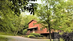 Une maison en bois rouge, dotée d’un porche et d’un escalier, entourée d’arbres et bordée d’un chemin de gravier.