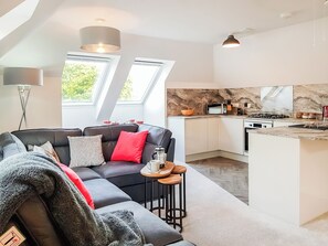 A modern apartment with a living area featuring a black leather sofa, a small round table, and a red pillow. The kitchen has white cabinets, a marble backsplash, and a built-in stove. There are skylights providing natural light. - Palace View - UK40569 (Falkland)