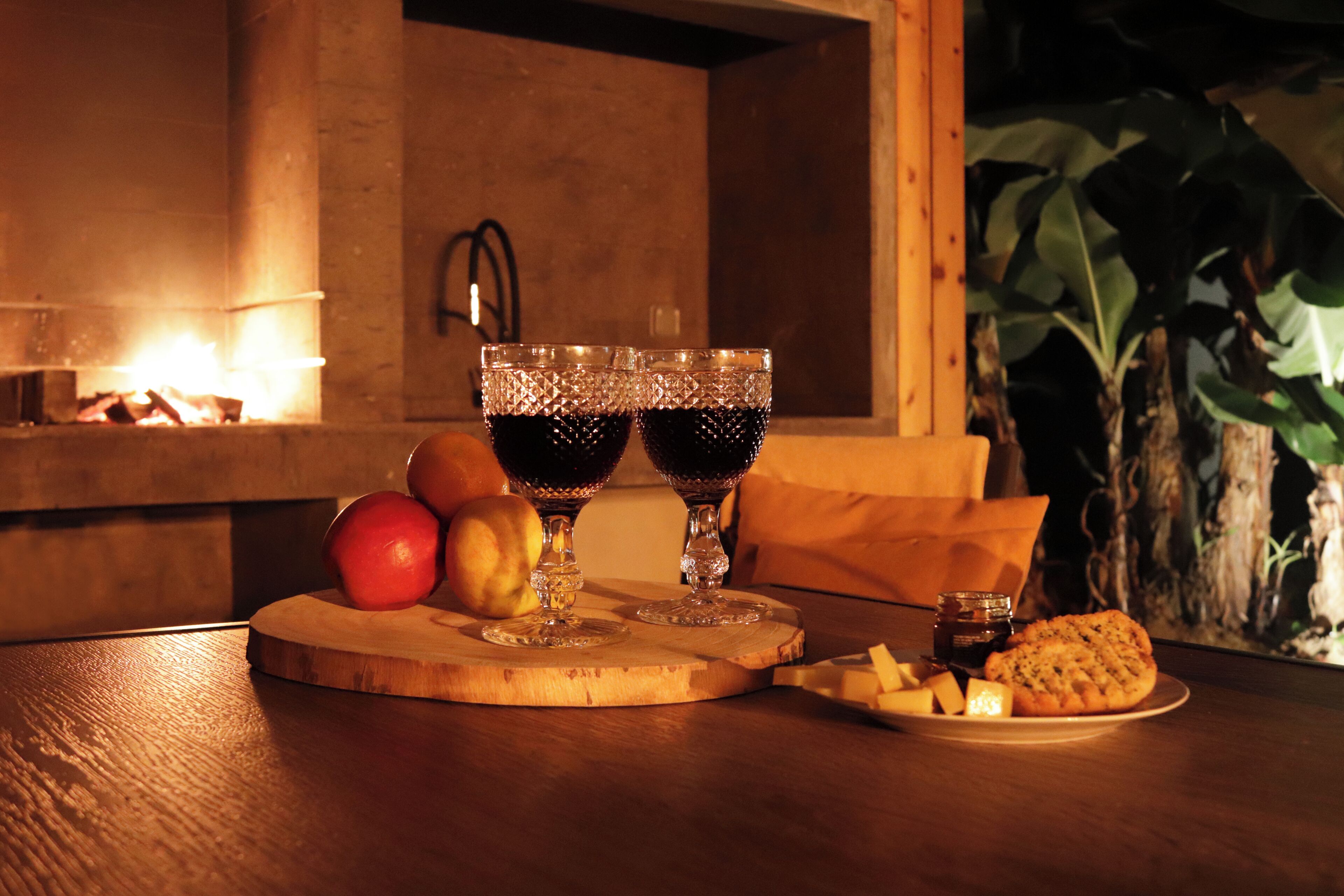 A wooden table with a plate of cheese and crackers, two glasses of red wine, and a bowl of fruit.