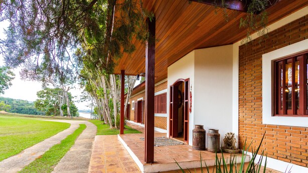 A covered outdoor area with a brick wall, wooden beams, and a pathway leading to a building entrance. - Sítio King - Bragança Paulista (Piracaia)