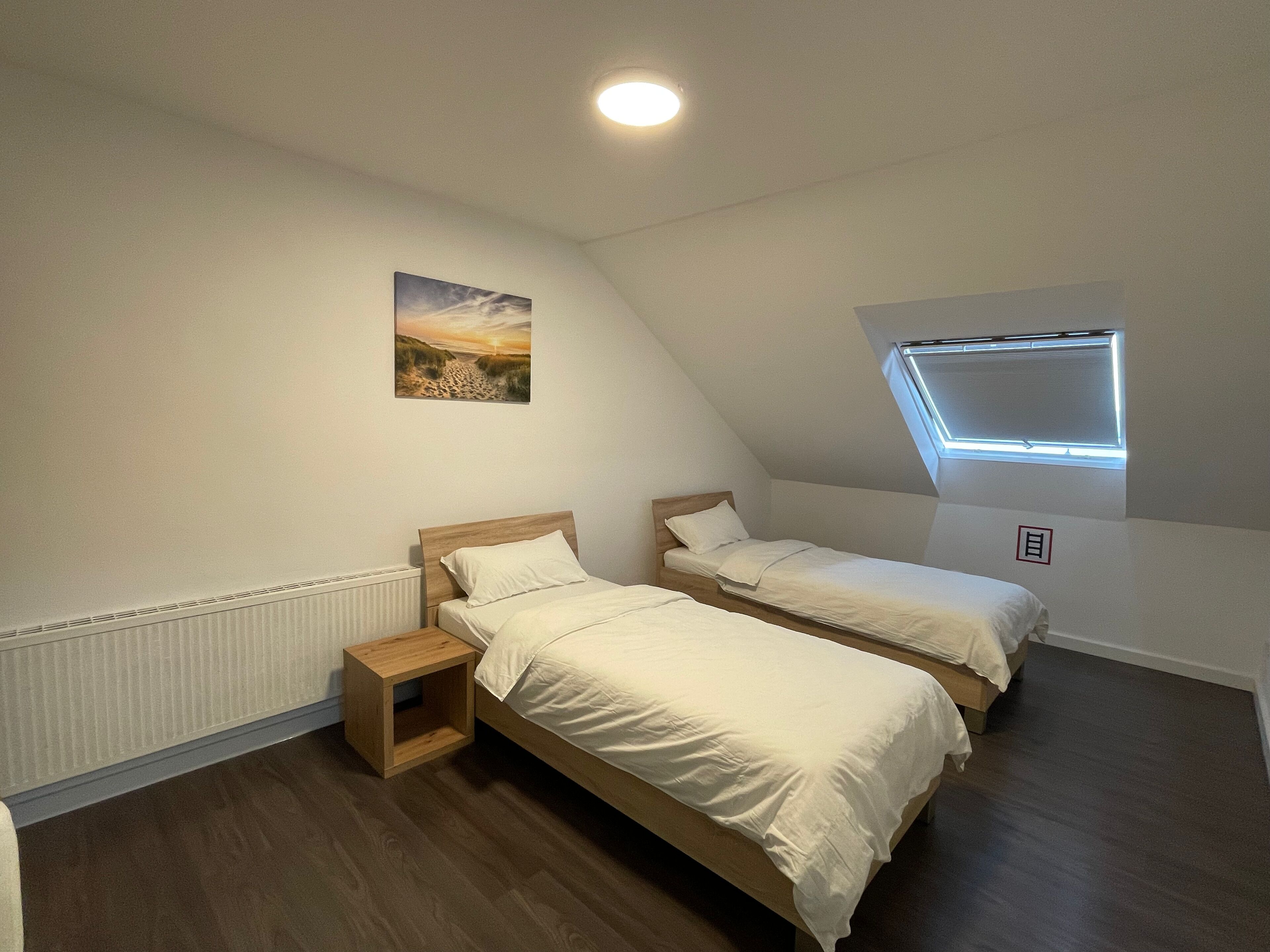 A bedroom with two single beds, a small bedside table, a radiator, a picture on the wall, and a skylight.
