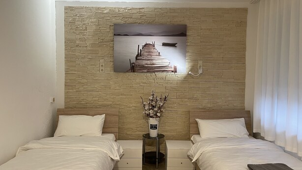 A hotel room with two beds, a stone wall, a painting of a dock, and a vase with flowers. - Erft Apartment (Kerpen)