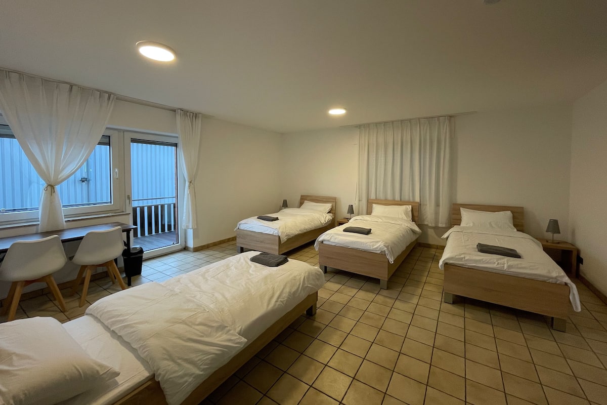 A hotel room with three single beds, a desk, chairs, and a balcony with curtains.