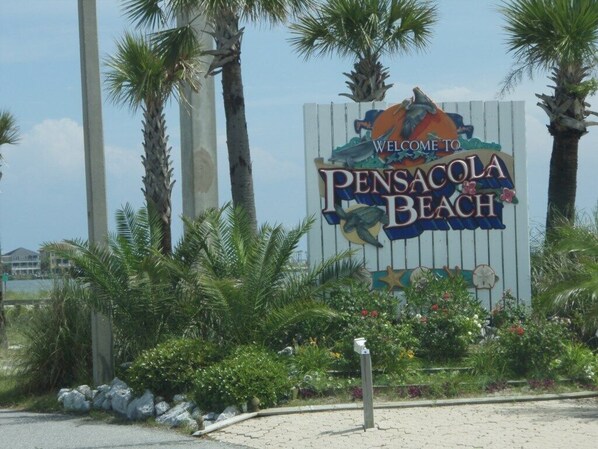 Señal de bienvenida a Pensacola Beach con palmeras y flores al frente.