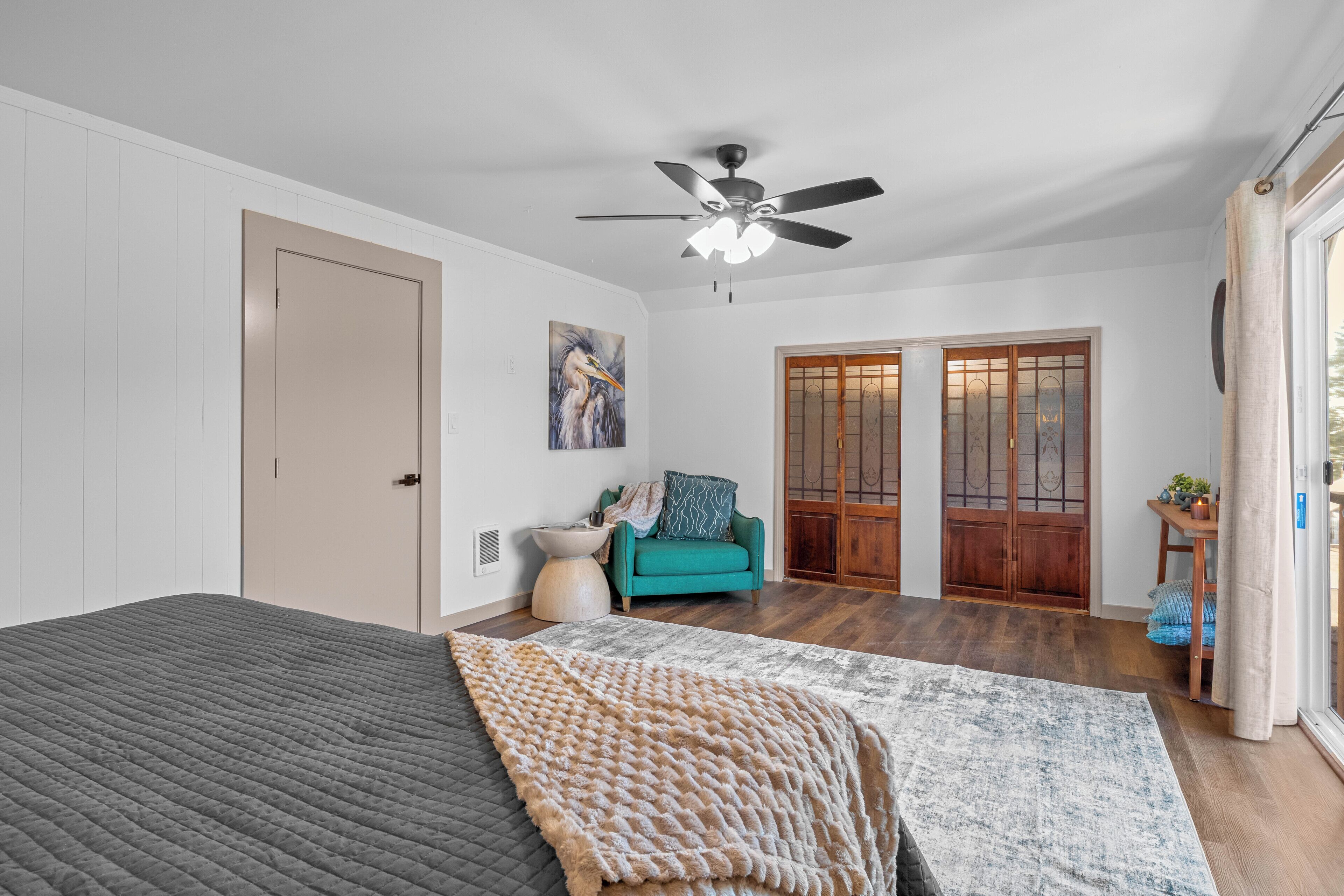 Une chambre à coucher avec un lit, un ventilateur de plafond, un tableau, une porte et une vue sur l’extérieur.