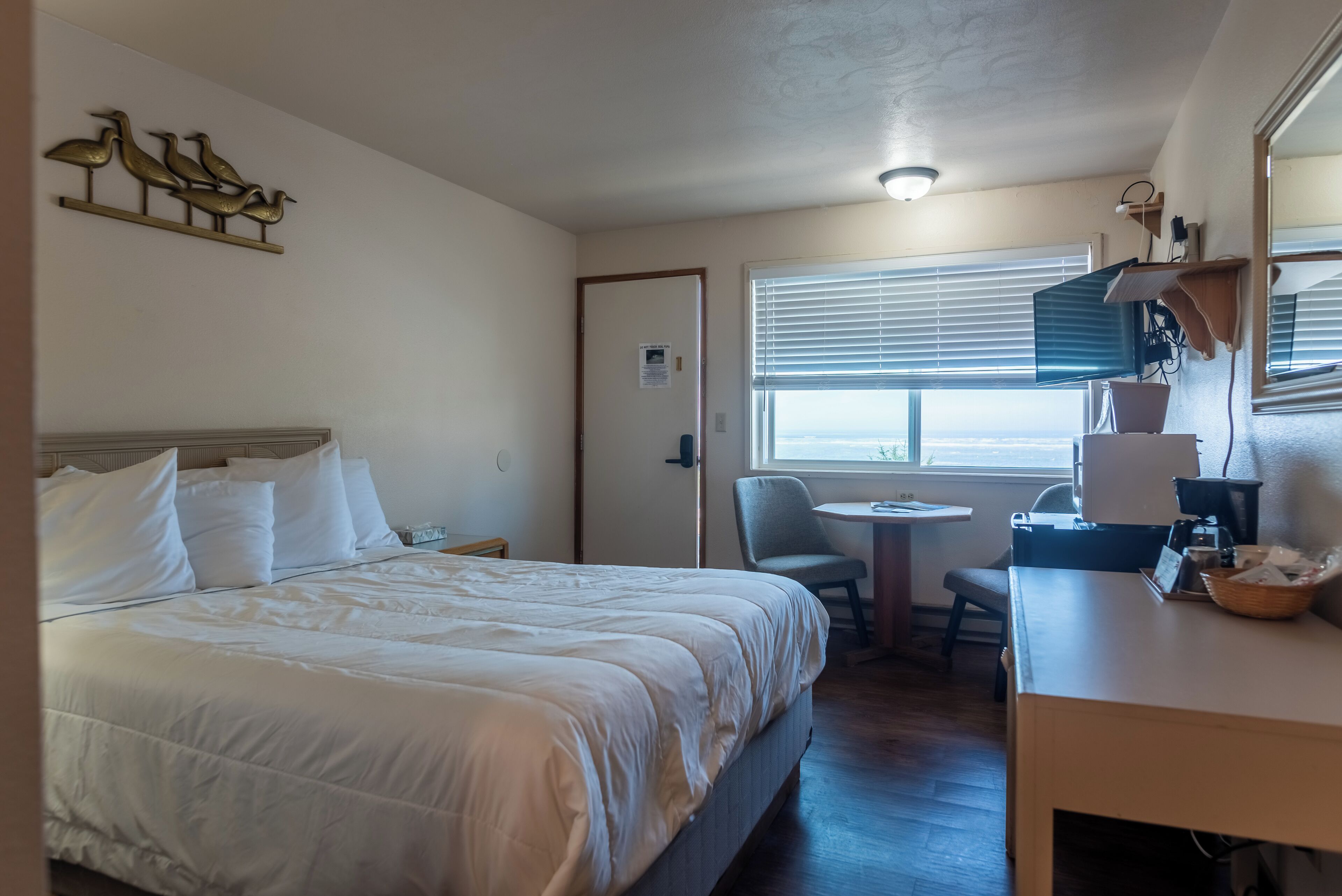 A hotel room with a bed, a desk, a chair, a window with blinds, and a wall-mounted shelf with decorative items.