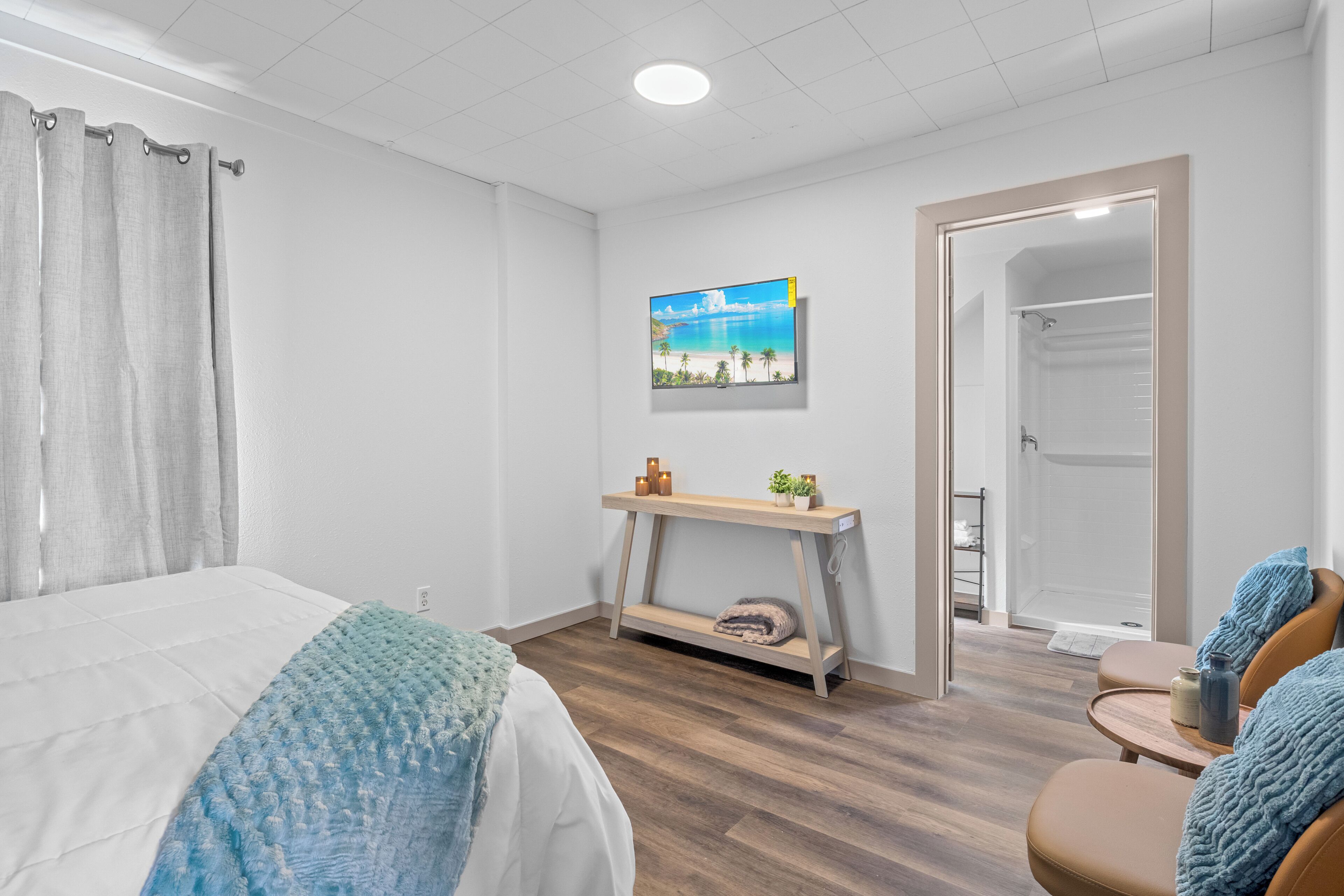 A hotel room with a bed, a small table with candles, a painting of a beach, and a shower area.