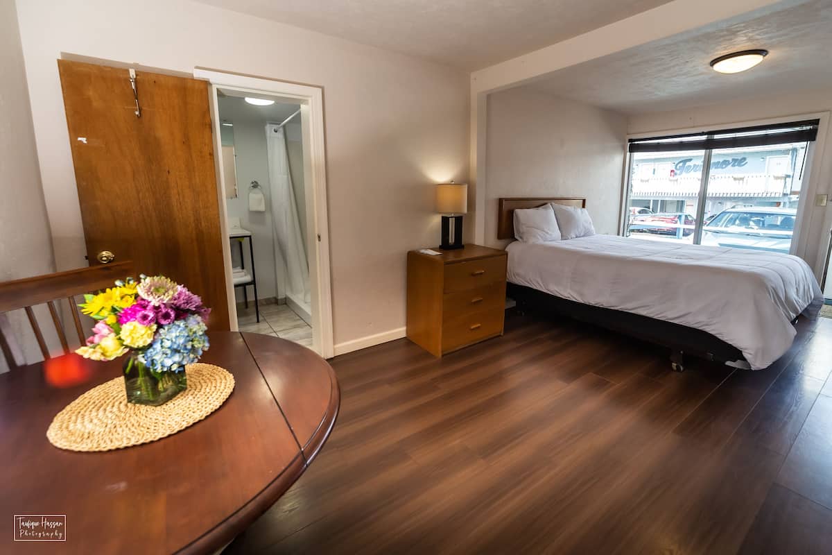 Ein Hotelzimmer mit Bett, Nachttisch mit Lampe, einem Holzbeistelltisch, einem Esstisch mit Blumenvase und Blick durch ein großes Fenster nach draußen.