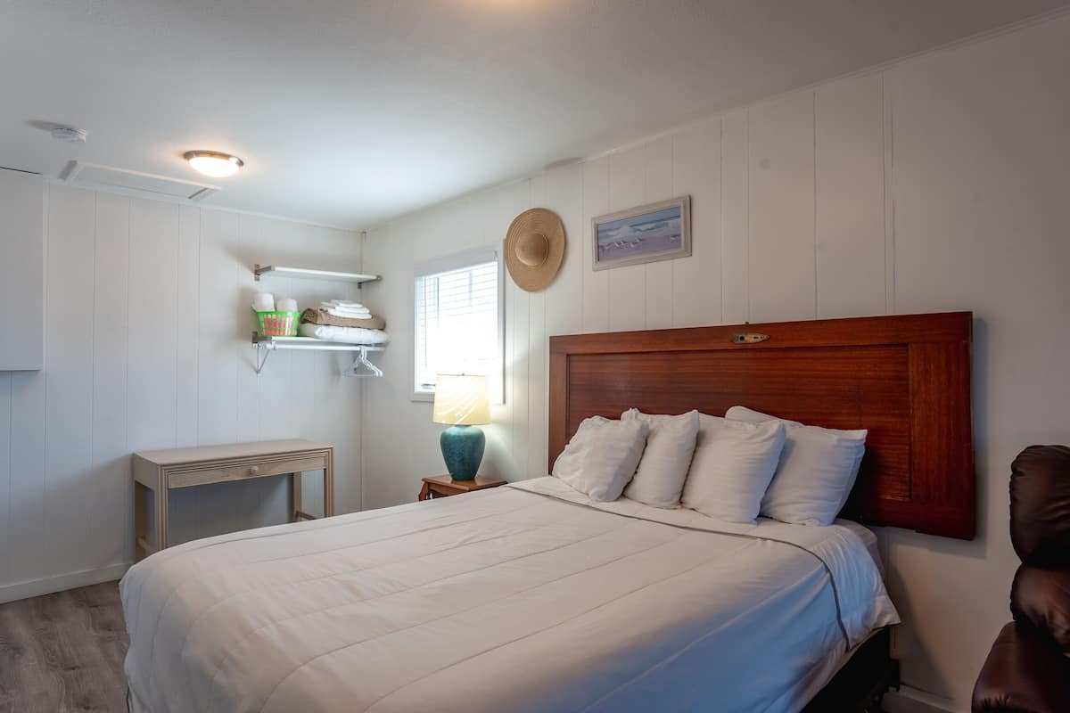 Ein Schlafzimmer mit einem großen Bett, einem Nachttisch mit Lampe, einem Holz-Kopfteil, einem Hut an der Wand und einem Regal mit Handtüchern.
