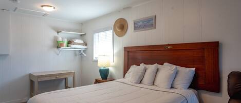 A bedroom with a large bed, a nightstand with a lamp, a wooden headboard, a hat on the wall, and a shelf with towels.