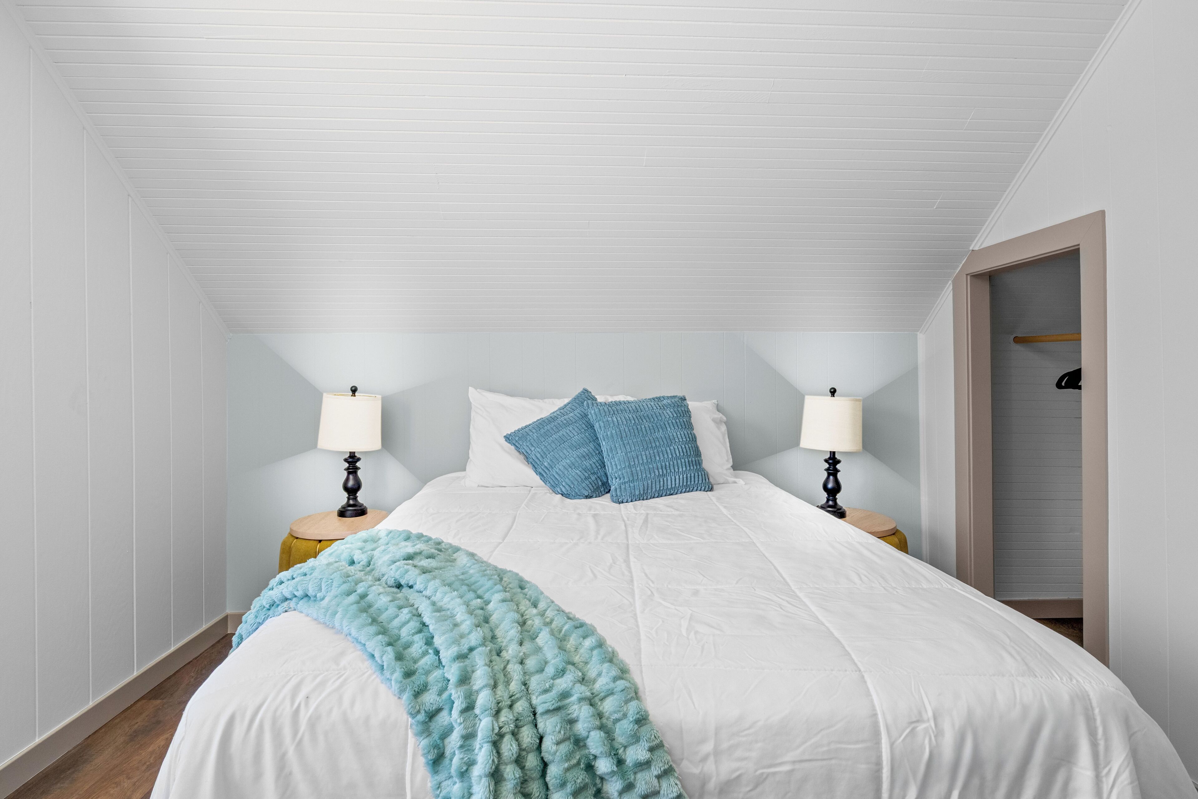 Un dormitorio con techo inclinado, una cama con ropa de cama blanca, dos mesitas de noche con lámparas y un armario con barra de madera.