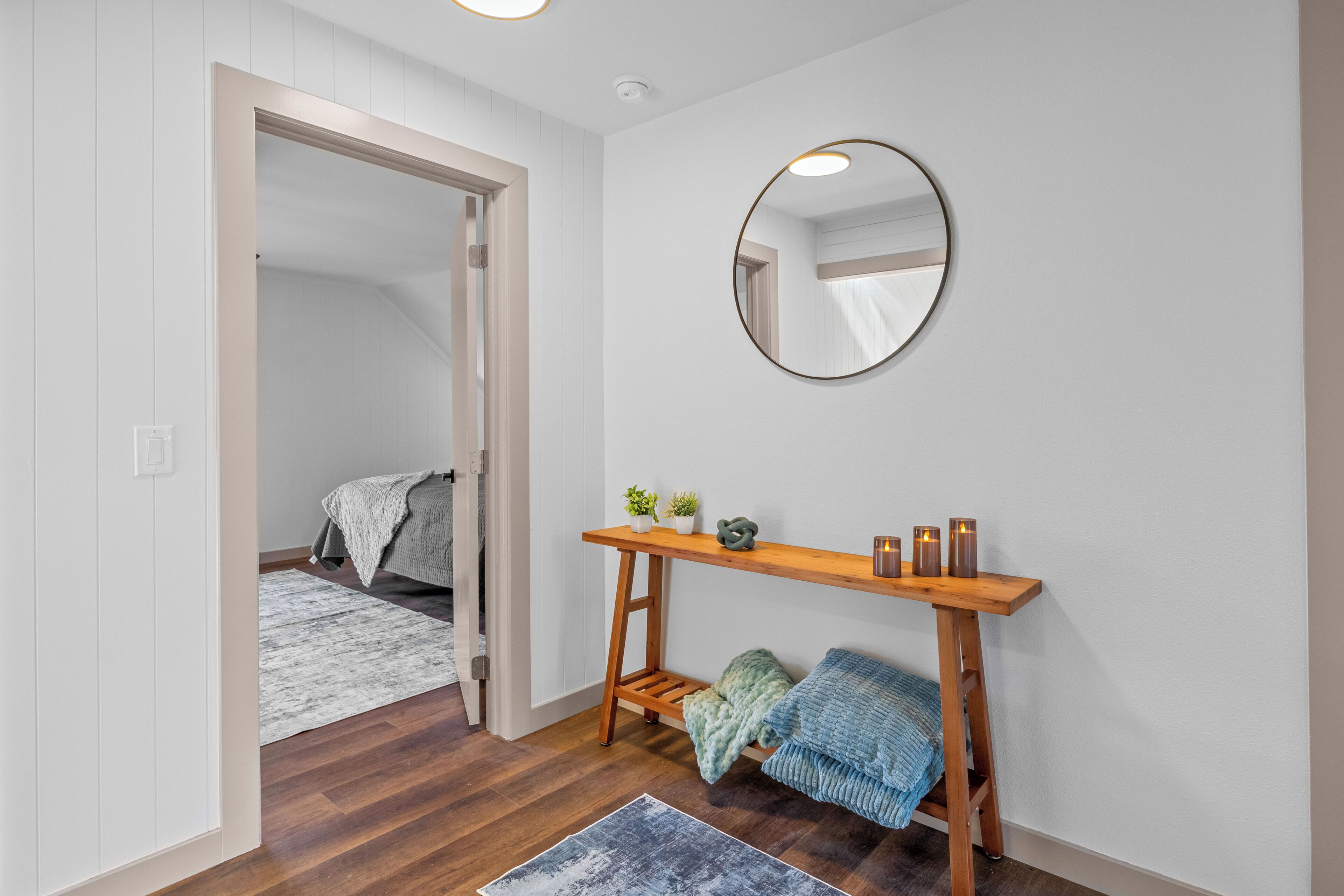 A room with a wooden console table, a round mirror, a bed with a grey blanket, and a small potted plant.
