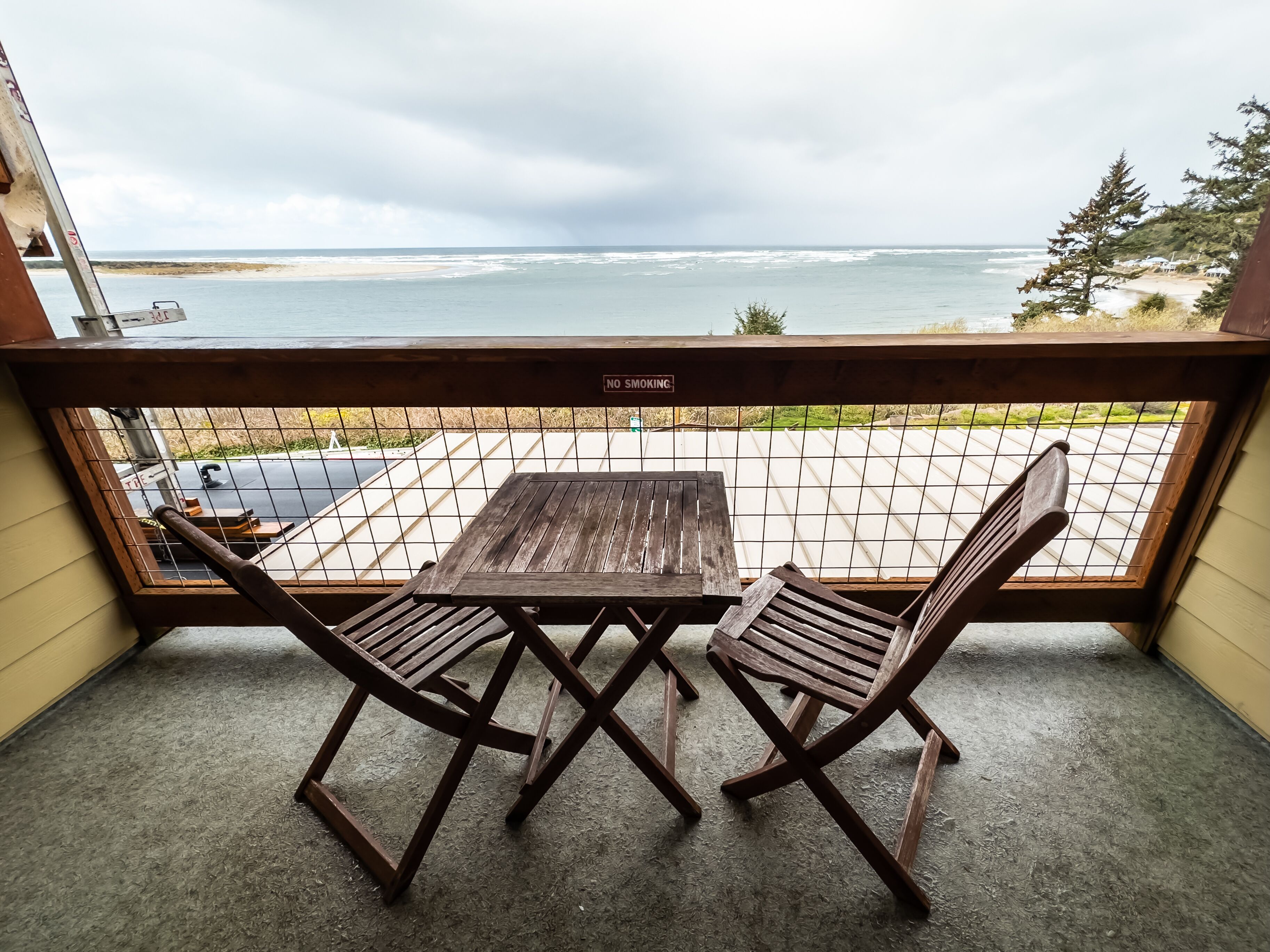 A balcony with a wooden table and two chairs, overlooking a beach and ocean.