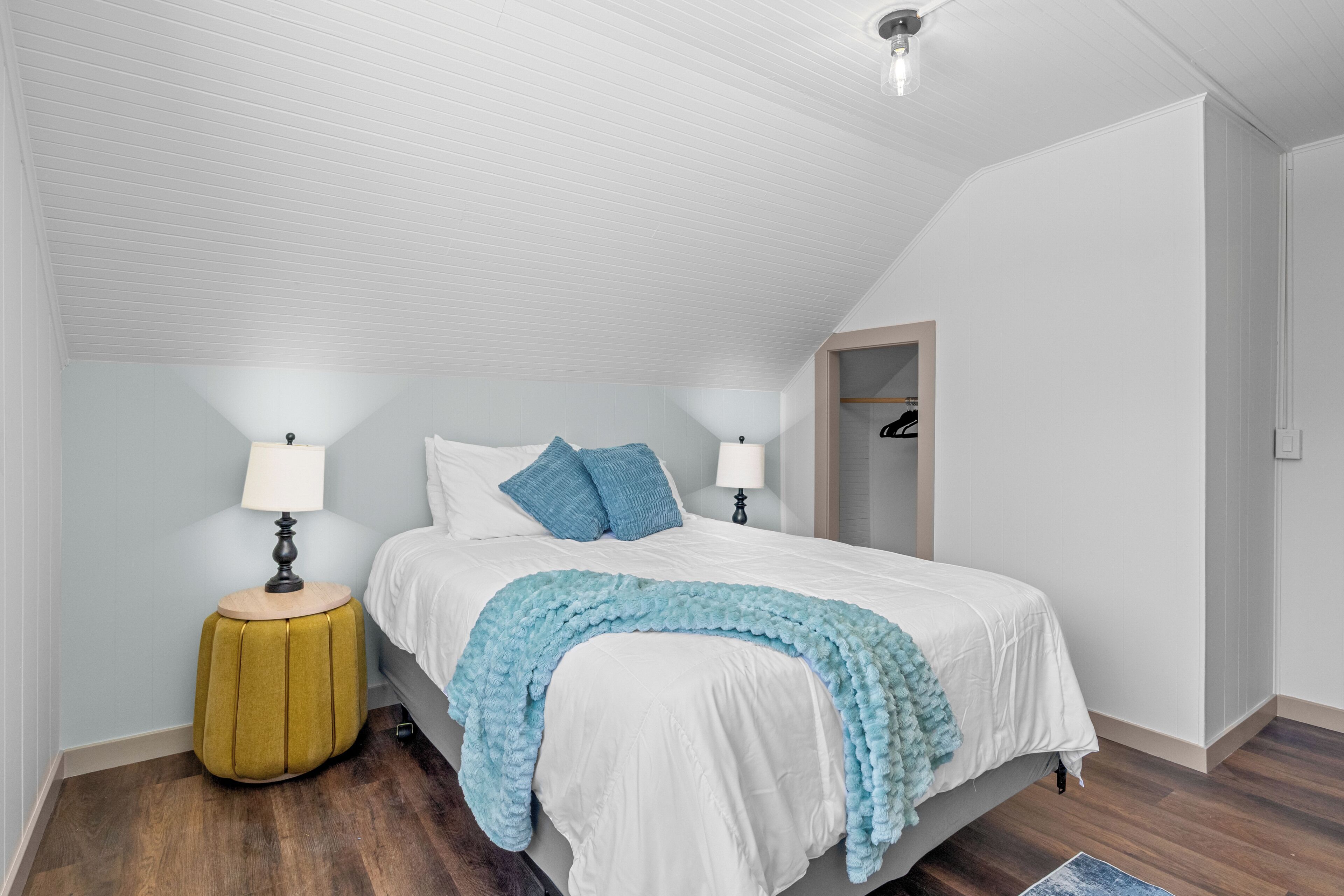 A bedroom with a bed, two bedside lamps, a nightstand, a wooden stool, and a door leading to a closet.