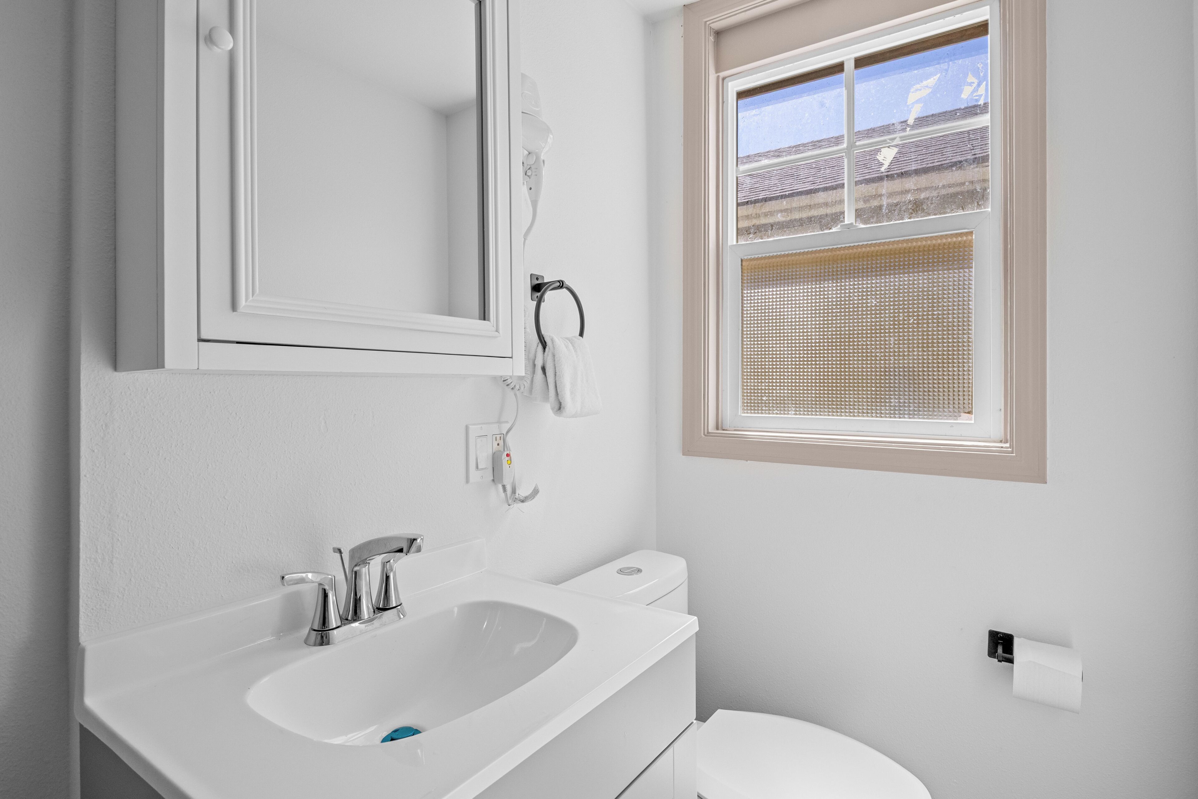 Un baño con lavamanos blanco, un espejo, un inodoro y una ventana con persianas.