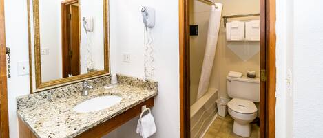 A bathroom with a granite countertop sink, a large mirror, a hairdryer, and a shower area.