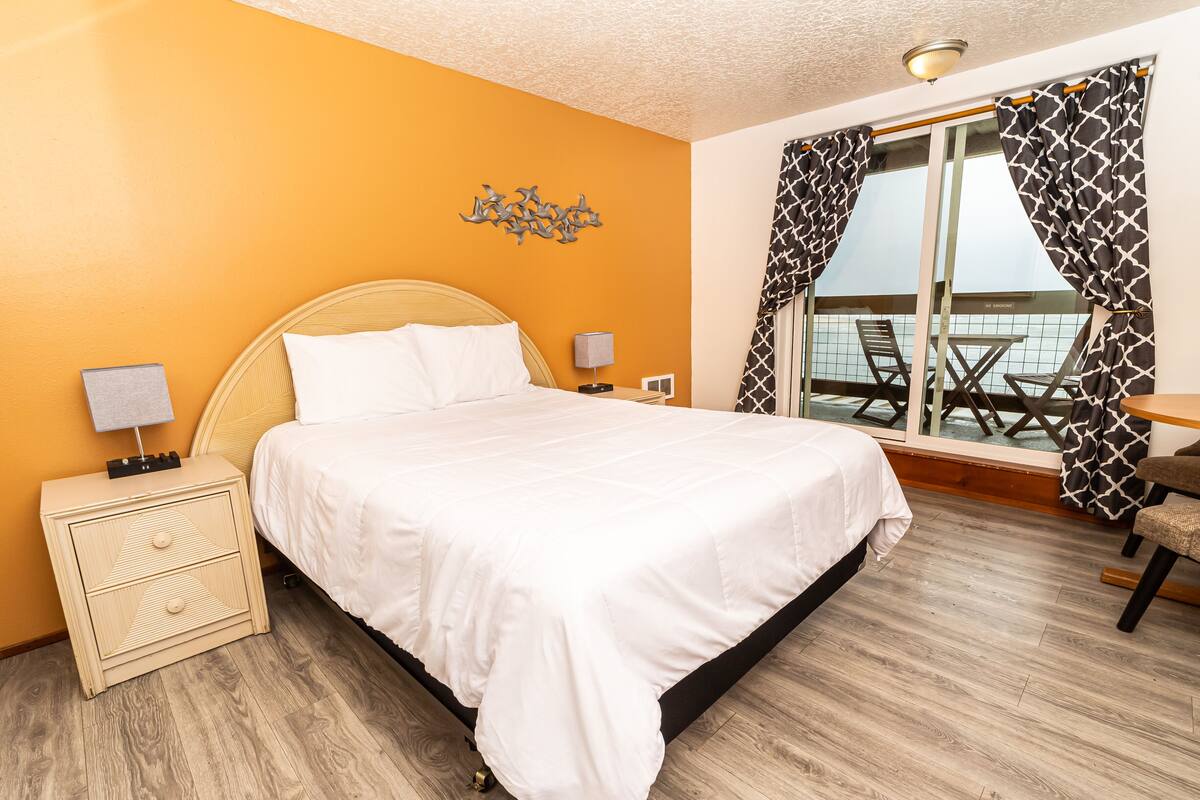 Ein Hotelzimmer mit Bett, Nachttischen, Lampe, Fenster mit Vorhängen und Balkon mit Tisch und Stühlen.