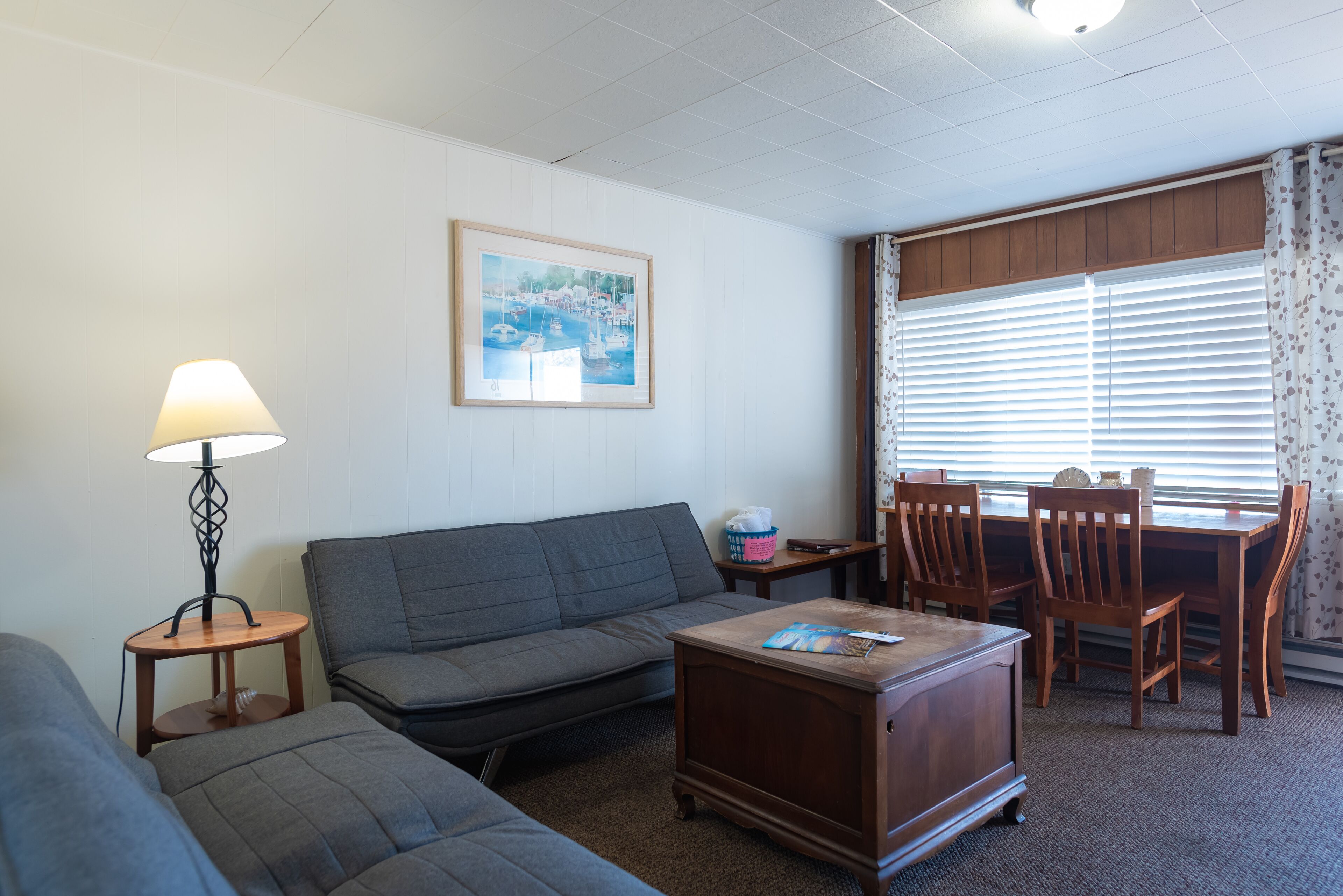 A living room with a sofa, a coffee table, a lamp, a framed picture on the wall, and a dining area with chairs and a table.