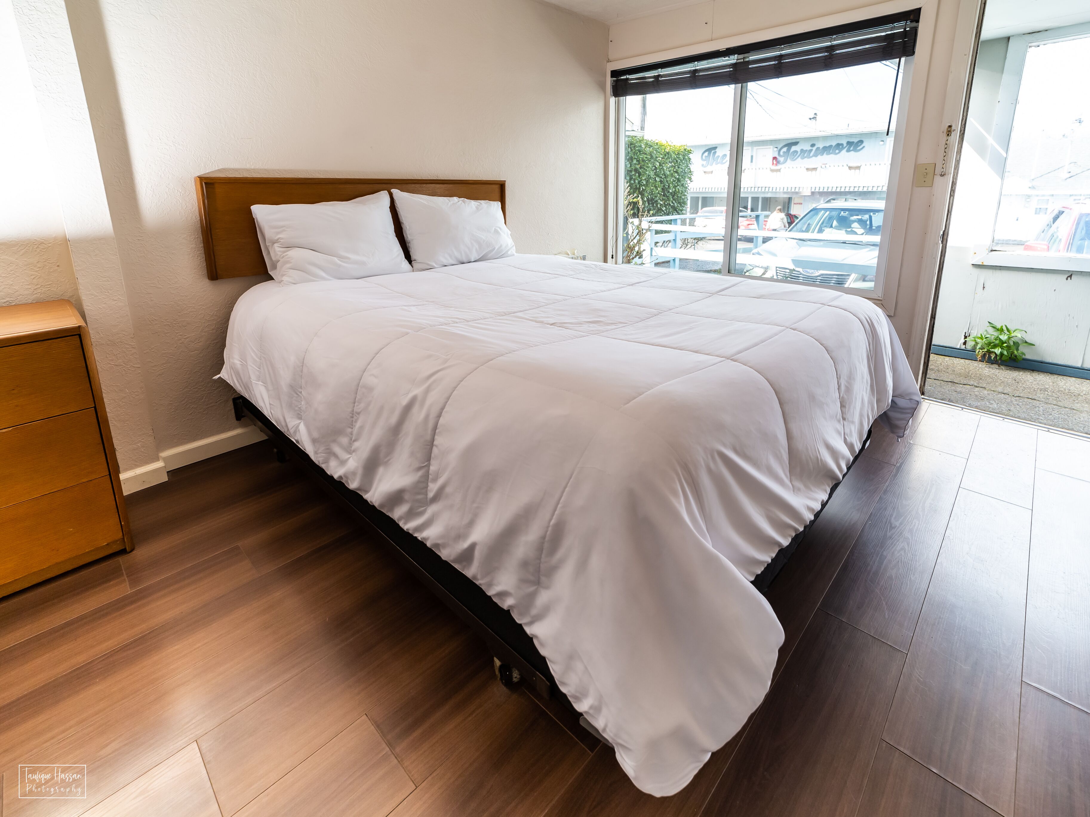 A neatly made bed in a room with a wooden headboard, a wooden nightstand, and a view of the street through a large window.