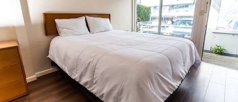 A neatly made bed in a room with a wooden headboard, a wooden nightstand, and a view of the street through a large window.