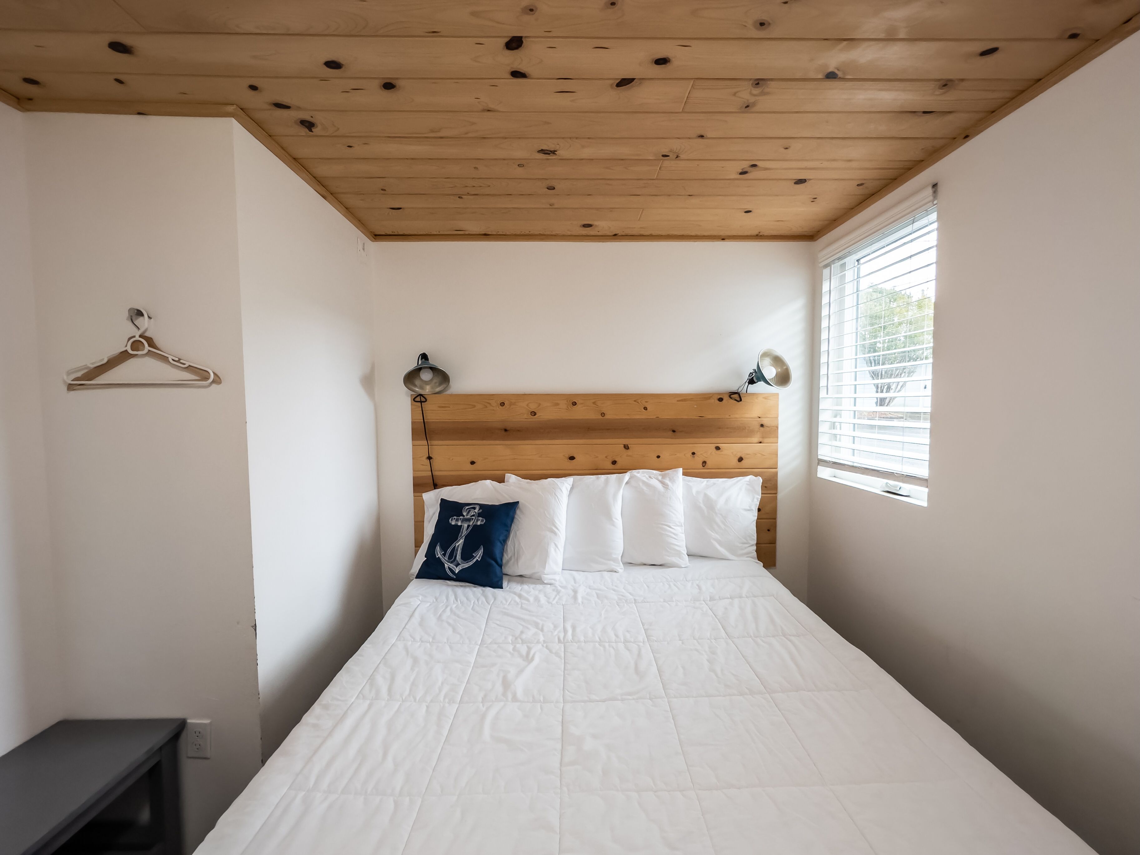 Een slaapkamer met een houten plafond, een bed met wit beddengoed en een blauw kussen met anker dessin, een bedlampje en een raam met zonwering.