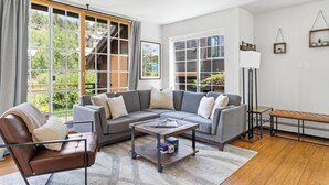 A modern living room with a sectional sofa, a leather armchair, a coffee table, and a bench. There are large windows with curtains, a floor lamp, and wall-mounted shelves. - Mountain Vista: Beautiful Basecamp @ Meadows Lift (Mountain Village)