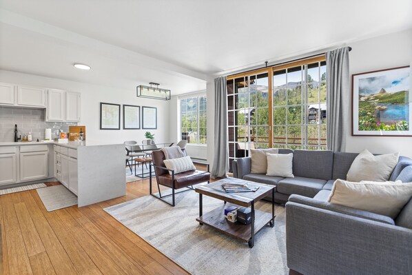 A modern living room with a grey sofa, a coffee table, and a view of a mountainous landscape through large windows.
