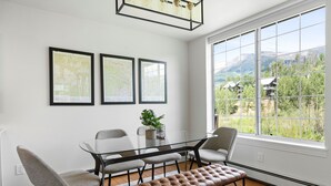 A modern dining area with a glass table, cushioned chairs, and a bench. There are three framed artworks on the wall and a large window offering a view of greenery and mountains. - Mountain Vista: Beautiful Basecamp @ Meadows Lift (Mountain Village)