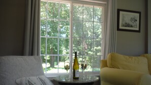 Una habitación con una ventana, un sillón amarillo, un sillón blanco y una mesita con una botella y un vaso.