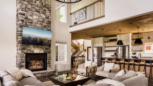 A spacious living area with a stone fireplace, a high ceiling, and a kitchen area with modern appliances. - Swim Spa/Luxe Design/Views/GameRoom/Theater - Bear Bottom (Gatlinburg)