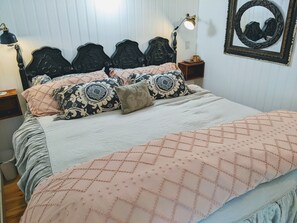 Een opgemaakt bed met gestreepte kussens en een gewatteerde spreidek, een nachtkastje met een lamp, en een spiegel aan de muur.