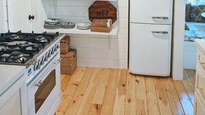 Une cuisine avec un réfrigérateur blanc, une cuisinière et des armoires en bois. Au-dessus du réfrigérateur, on peut lire une pancarte indiquant : « Épicerie et produits secs ».