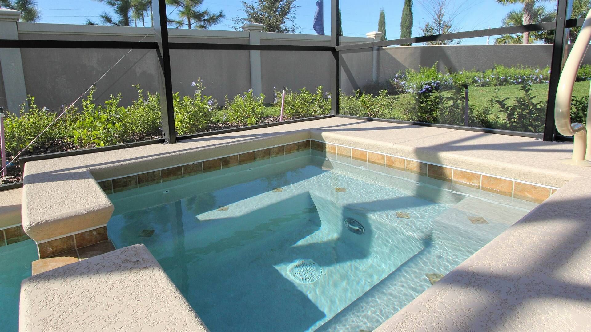 Una bañera de hidromasaje rectangular con borde de azulejos, rodeada por una terraza color beige y una valla metálica.