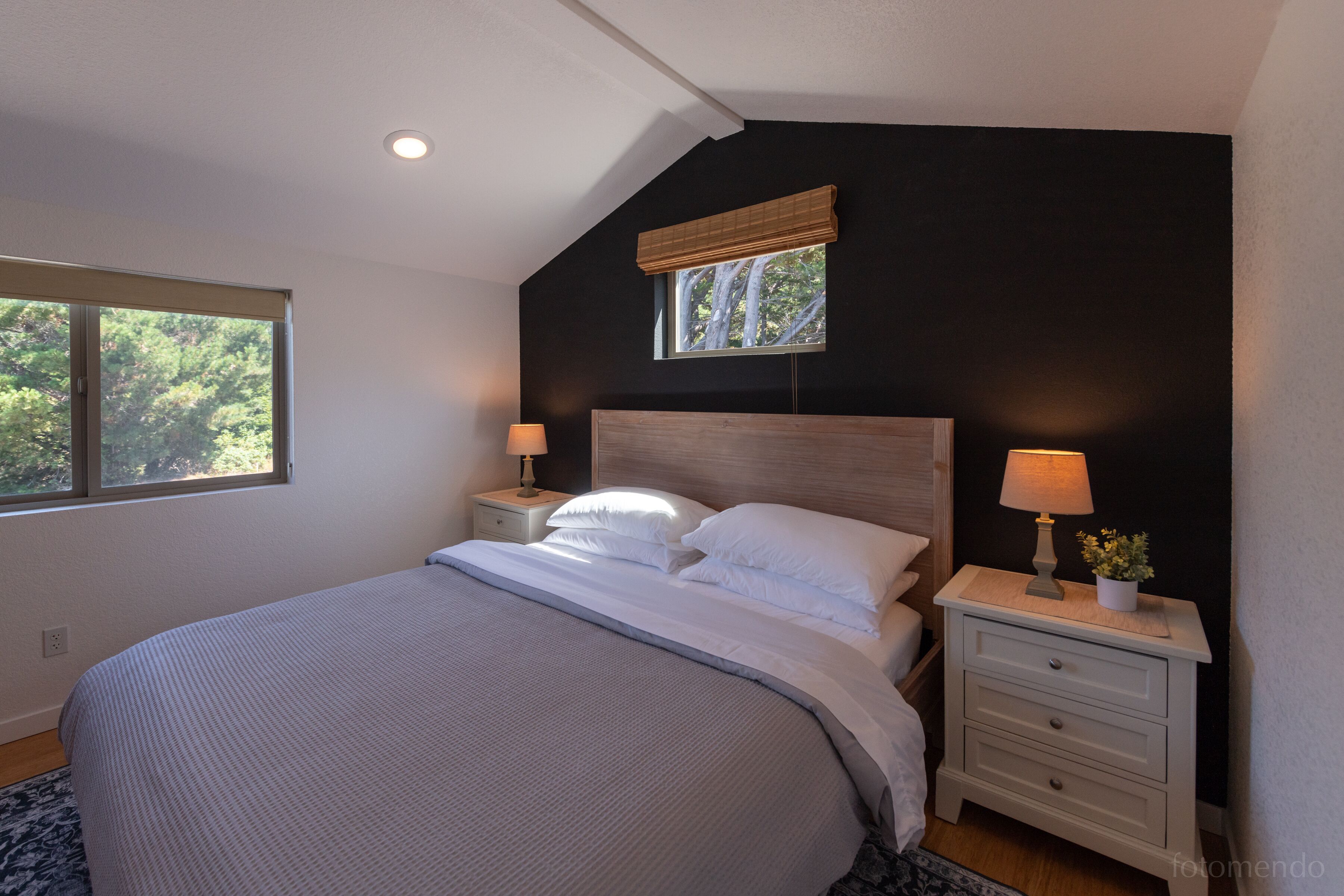 A bedroom with a large bed, two bedside tables with lamps, a window with a view of trees, and a dark accent wall.