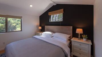 A bedroom with a large bed, two bedside tables with lamps, a window with a view of trees, and a dark accent wall.