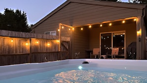 Un bain à remous agrémenté de guirlandes lumineuses, entouré d’une clôture en bois et doté d’un patio couvert.