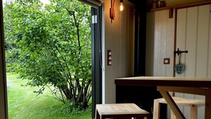 Una habitación con una mesa y banco de madera, vista a un jardín verde y una lámpara en la pared.