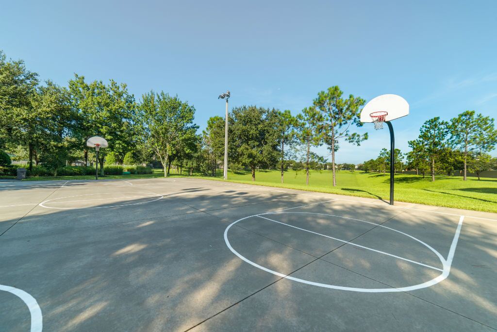 Quadra de basquete ao ar livre com duas aros, cercada por árvores e céu claro.