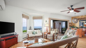 A spacious living room with a large flat-screen TV, wicker furniture, and a view of the ocean. - Aloha Condos, Kanaloa at Kona, Condo 1901, Oceanfront, AC (Kailua Kona)