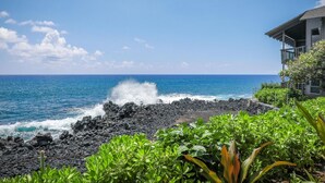 A coastal view with a rocky shoreline, ocean waves, and a building with a balcony overlooking the sea. - Aloha Condos, Kanaloa at Kona, Condo 1901, Oceanfront, AC (Kailua Kona)