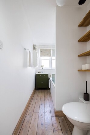 Een smalle badkamer met een houten vloer, een groen kastje, een wit toilet en een raam met uitzicht.