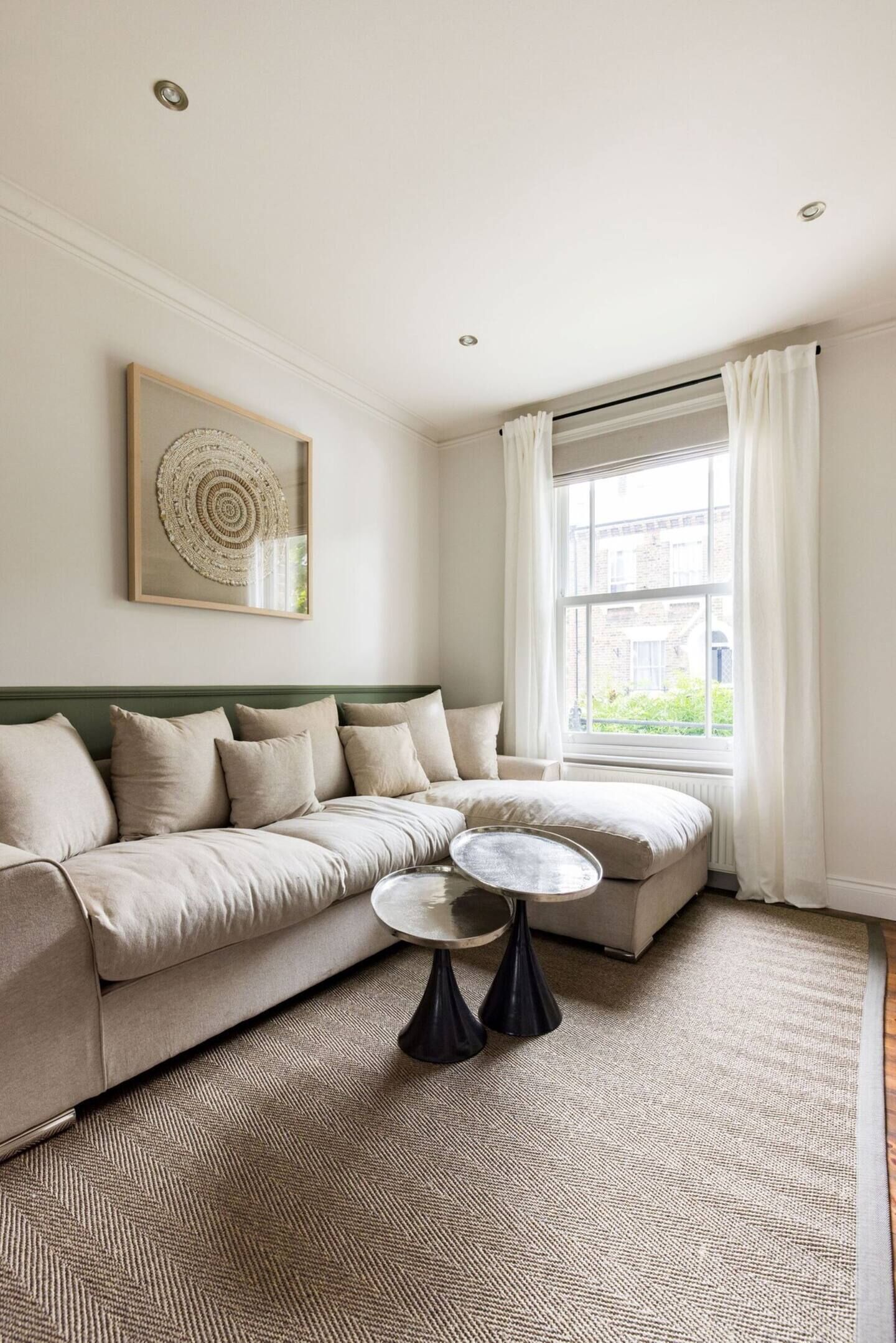 Ein Wohnzimmer mit einem beigefarbenen Ecksofa, einem runden Couchtisch und einem großen Fenster mit weißen Vorhängen.