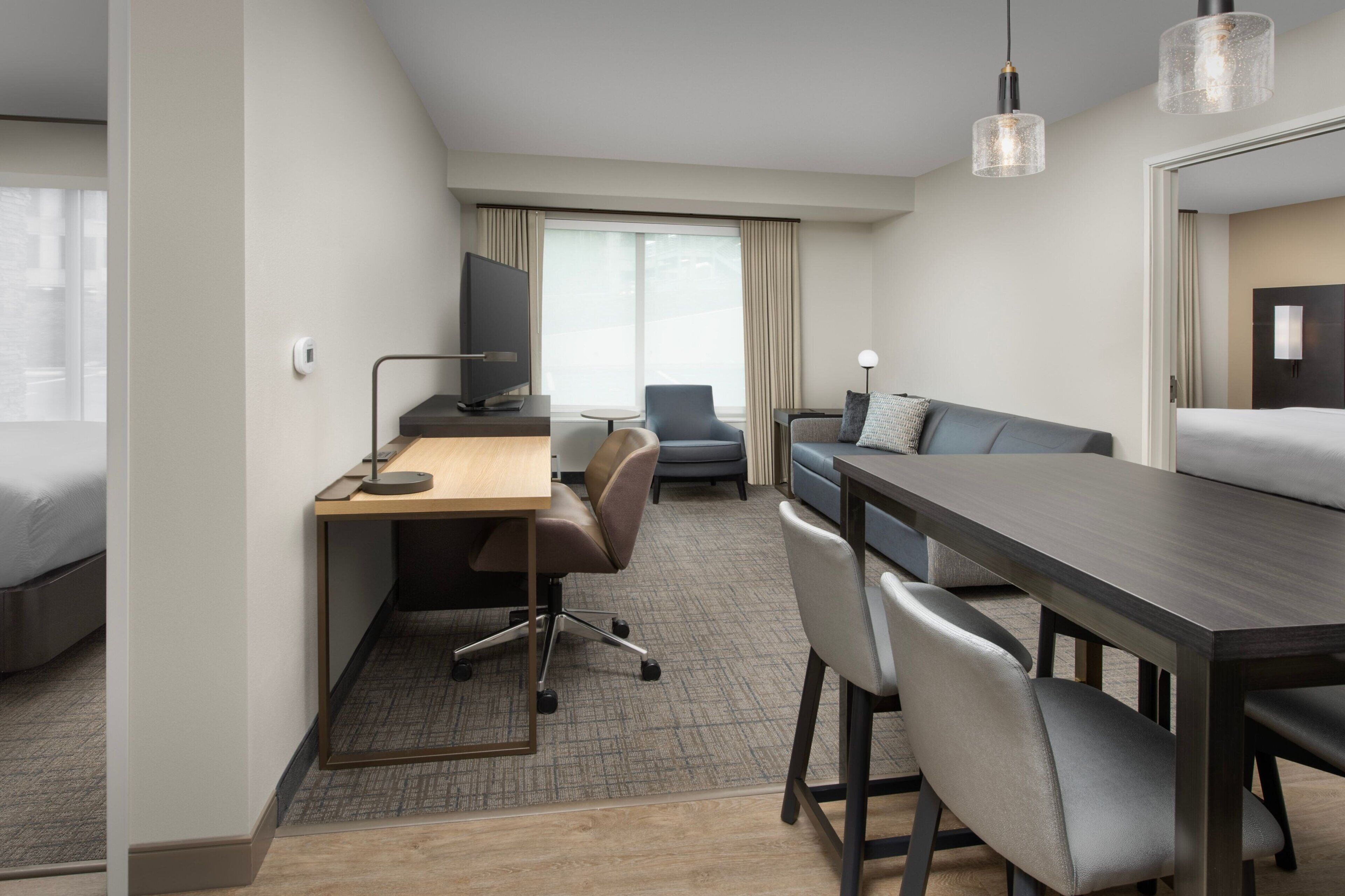 A modern hotel room with a desk, two chairs, a sofa, a TV, and a large window with curtains.