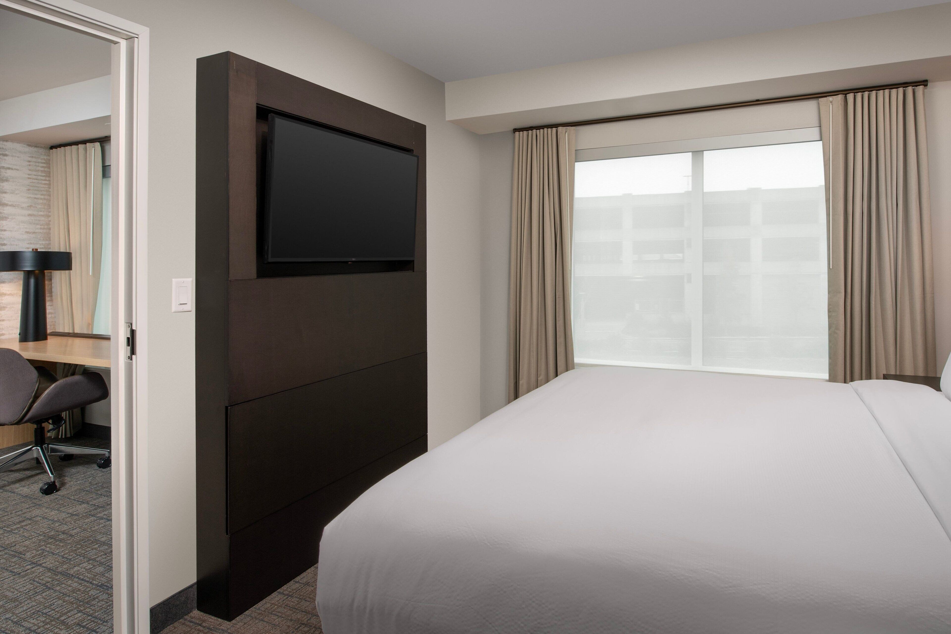 A hotel room with a large bed, a flat-screen TV mounted on the wall, a desk with a chair, and a window with curtains.