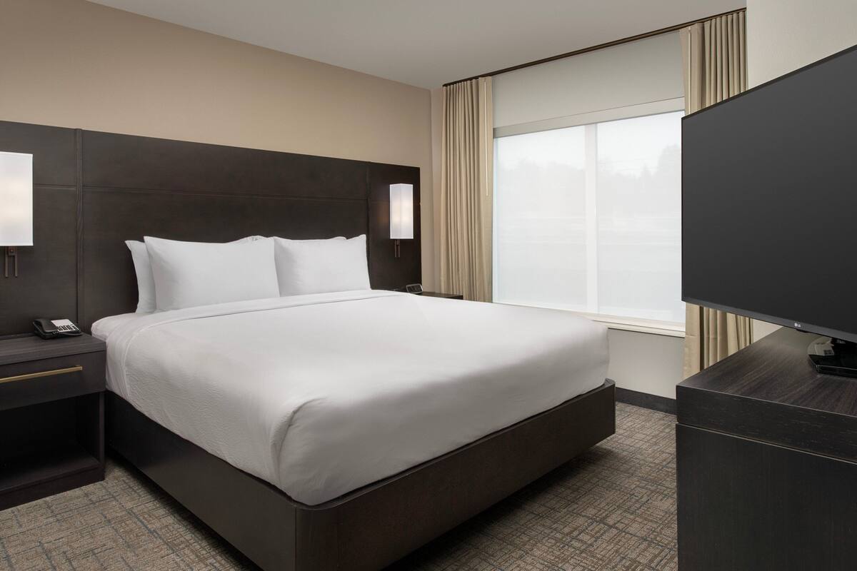 Une chambre d’hôtel avec un grand lit, une table de chevet avec une lampe, une télévision et une fenêtre avec des rideaux.